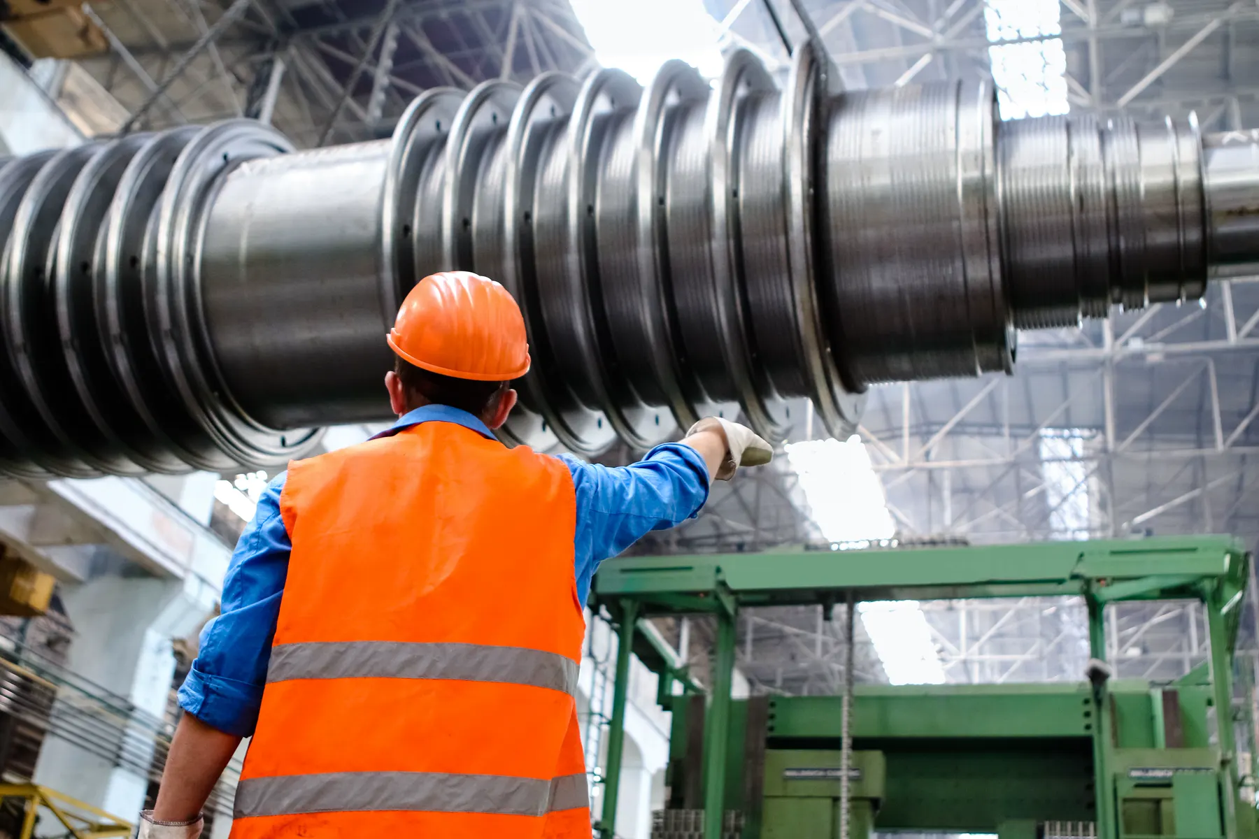 Worker in hi-vis, seen from behind, looking at a large machined shaft on a plant floor